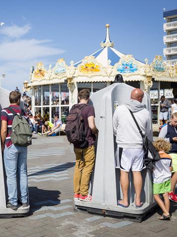 Op de scheveningse boulevard staan urinoirs waar 3 mannen aan het plassen zijn. Daarnaast lopen kindjes. 