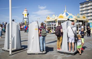 Op de scheveningse boulevard staan urinoirs waar 3 mannen aan het plassen zijn. Daarnaast lopen kindjes.