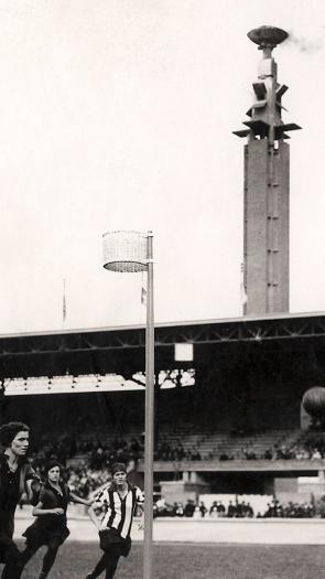 Zwart wit foto van vrouwenhandbal wedstrijd in het Olympisch Stadion in Amsterdam met op de achtergrond de marathontoren waar de olympische vlam rookt