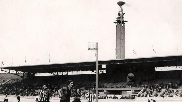 Zwart wit foto van vrouwenhandbal wedstrijd in het Olympisch Stadion in Amsterdam met op de achtergrond de marathontoren waar de olympische vlam rookt