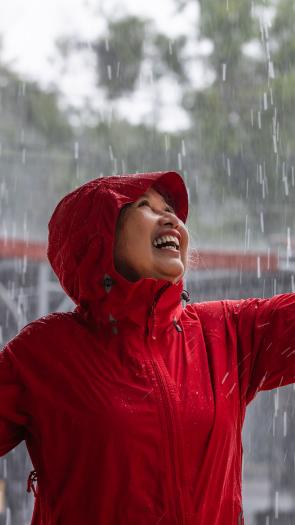 Een vrouw met regenjas staat in de regen met haar hand uitgestrekt en een lach op haar gezicht