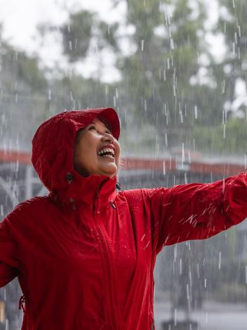 Een vrouw met regenjas staat in de regen met haar hand uitgestrekt en een lach op haar gezicht