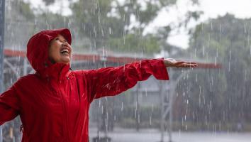 Een vrouw met regenjas staat in de regen met haar hand uitgestrekt en een lach op haar gezicht