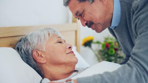 Een vrouw ligt ziek in bed; haar man buigt over haar heen en kijkt haar aan