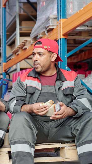 twee mannen zitten samen op een palet in een magazijn in werkkleding pauze te houden met eten en drinken