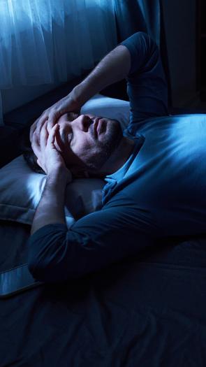 Man ligt in het donker in bed met handen tegen zijn hoofd aan en moeilijke blik