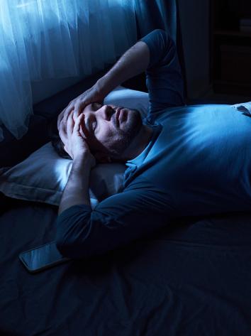 Man ligt in het donker in bed met handen tegen zijn hoofd aan en moeilijke blik