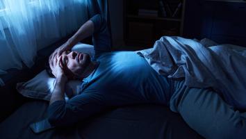 Man ligt in het donker in bed met handen tegen zijn hoofd aan en moeilijke blik