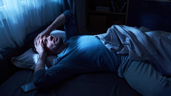 Man ligt in het donker in bed met handen tegen zijn hoofd aan en moeilijke blik