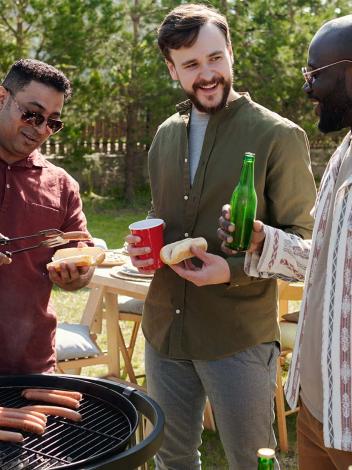 Drie mannen staan naast een barbecue broodjes met hotdogs te maken en te drinken en te kletsen