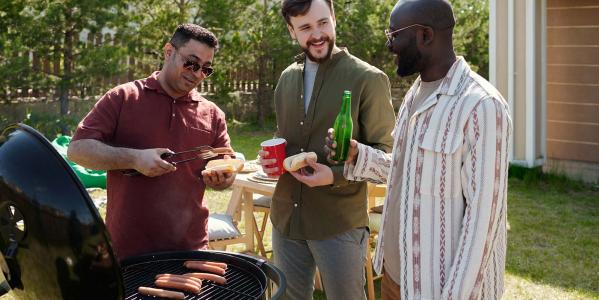 Drie mannen staan naast een barbecue broodjes met hotdogs te maken en te drinken en te kletsen