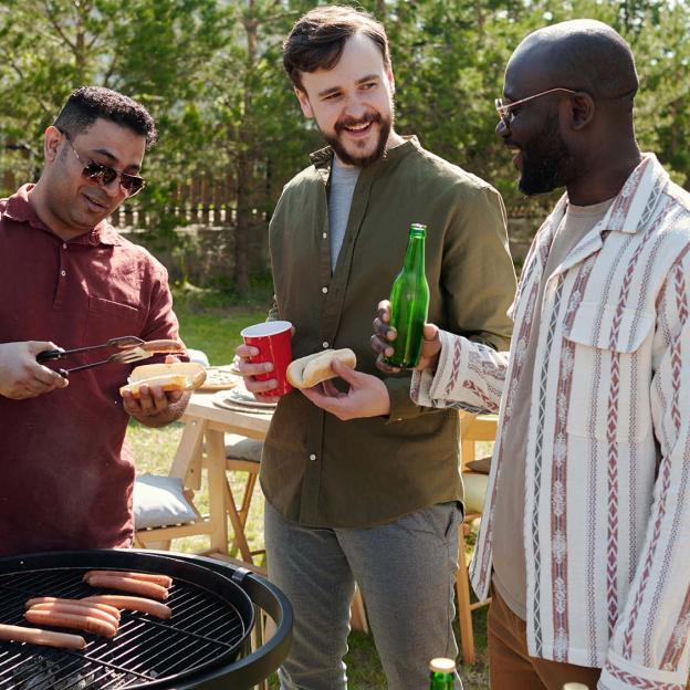 Drie mannen staan naast een barbecue broodjes met hotdogs te maken en te drinken en te kletsen