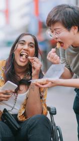 Iemand in een rolstoel en iemand die ernaast staat proeven samen kipnuggets uit een bakje 