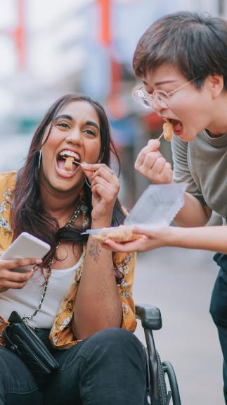 Iemand in een rolstoel en iemand die ernaast staat proeven samen kipnuggets uit een bakje 