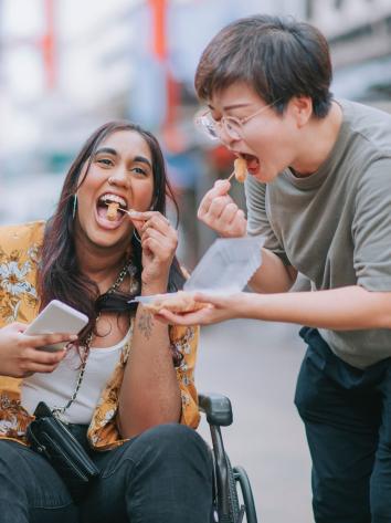 Iemand in een rolstoel en iemand die ernaast staat proeven samen kipnuggets uit een bakje 