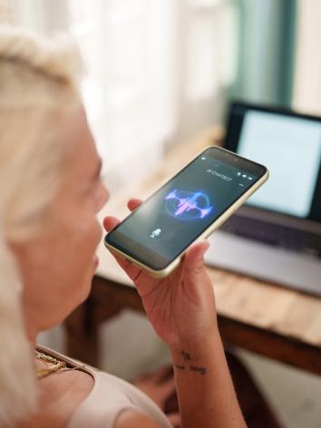 Een vrouw houdt haar telefoon vast en spreekt via de microfoon met een AI-chatbot. Op de achtergrond van de foto staat een laptop op een tafel