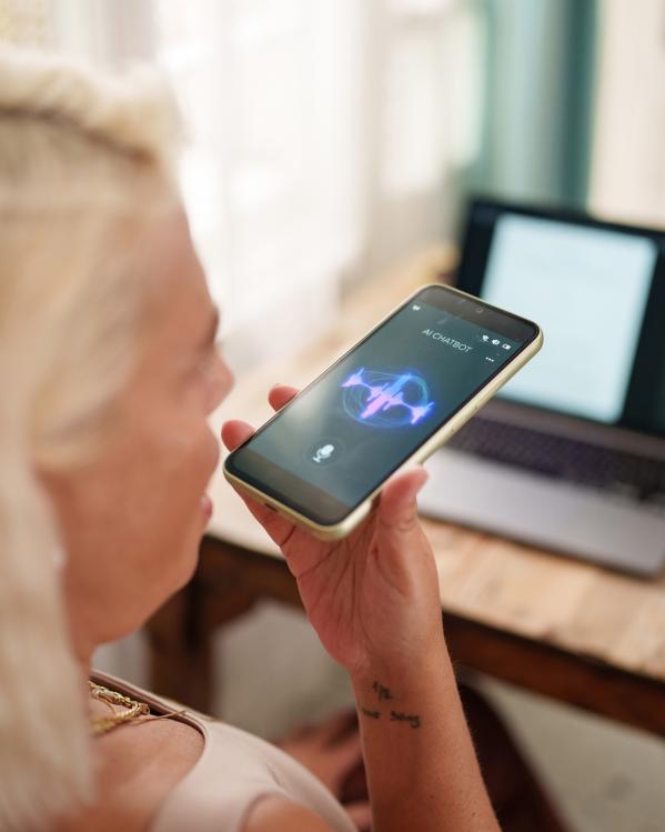 Een vrouw houdt haar telefoon vast en spreekt via de microfoon met een AI-chatbot. Op de achtergrond van de foto staat een laptop op een tafel