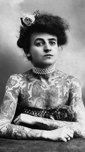 een zwart-witportretfoto van circusartiest Maud Wagner, wiens bovenlichaam vol tattoos zit van allerlei dieren en planten