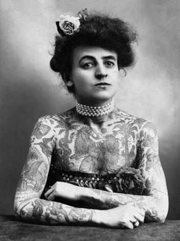 een zwart-witportretfoto van circusartiest Maud Wagner, wiens bovenlichaam vol tattoos zit van allerlei dieren en planten