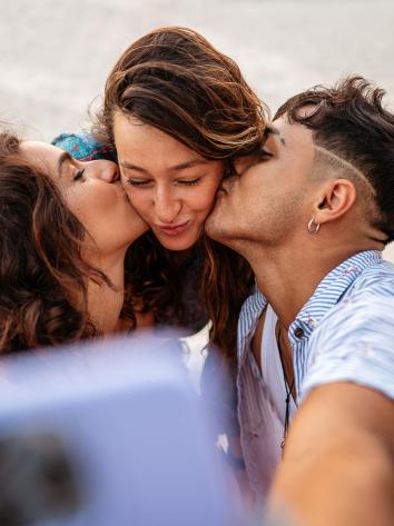 drie mensen maken een selfie waarbij de twee mensen aan de zijkant degene in het midden op de wang zoenen