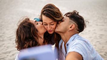 drie mensen maken een selfie waarbij de twee mensen aan de zijkant degene in het midden op de wang zoenen