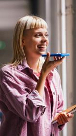 Iemand spreekt lachend iets in in haar telefoon, op de achtergrond staan andere mensen te praten