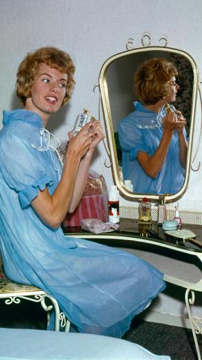 Zangeres Ria Valk (1941) in een blauwe nachtjapon achter haar kaptafel met spiegel, met een flesje parfum in haar handen, Nederland 1962.