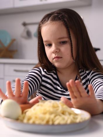Een jong meisje duwt het bord met pasta en ei weg met haar handen en kijkt afkeurend naar het eten