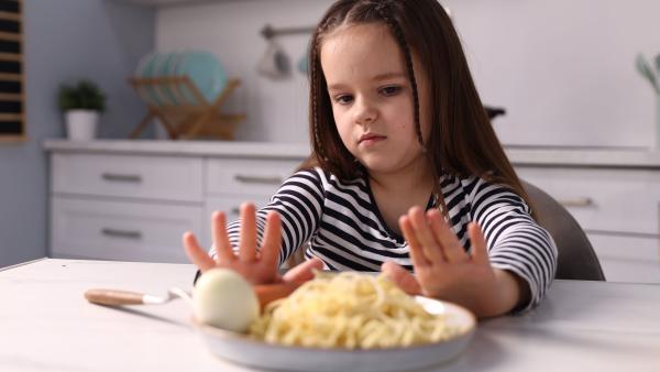 Een jong meisje duwt het bord met pasta en ei weg met haar handen en kijkt afkeurend naar het eten