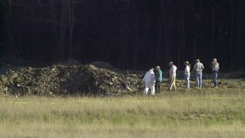 een aantal mensen doen onderzoek naar een grote krater waar het vliegtuig in Pennsylvania is neergestort, in een open grasveld met bos op de achtergrond.