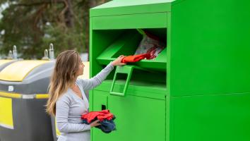 Vrouw stopt losse kledingstukken in de klep van een grote textielbak. 