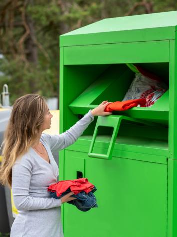 Vrouw stopt losse kledingstukken in de klep van een grote textielbak. 