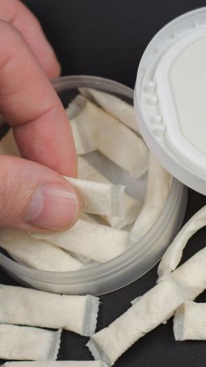 Een hand pakt een zakje snus uit een snusdoosje. Er liggen meer zakjes omheen.