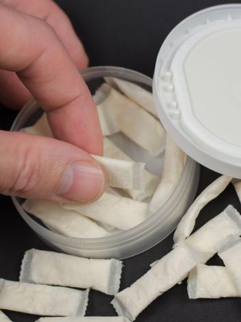 Een hand pakt een zakje snus uit een snusdoosje. Er liggen meer zakjes omheen.