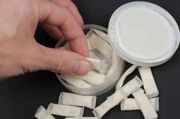 Een hand pakt een zakje snus uit een snusdoosje. Er liggen meer zakjes omheen.