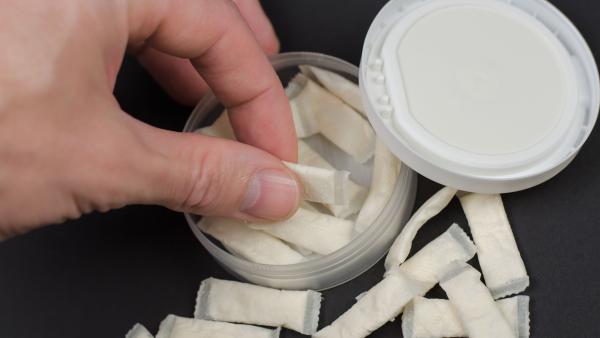 Een hand pakt een zakje snus uit een snusdoosje. Er liggen meer zakjes omheen.