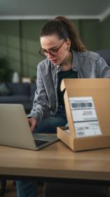 Vrouw met bril zit achter eettafel en typt iets op haar laptop terwijl ze een pakketje half open heeft