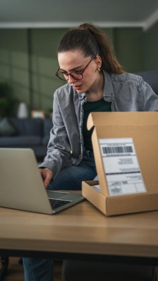 Vrouw met bril zit achter eettafel en typt iets op haar laptop terwijl ze een pakketje half open heeft