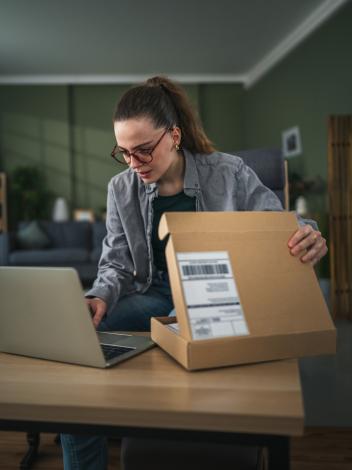 Vrouw met bril zit achter eettafel en typt iets op haar laptop terwijl ze een pakketje half open heeft