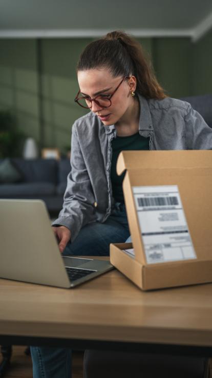 Vrouw met bril zit achter eettafel en typt iets op haar laptop terwijl ze een pakketje half open heeft