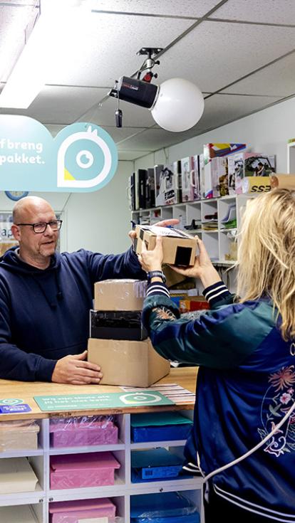 Vrouw geeft pakketjes af bij een postkantoor aan een man die achter de balie staat.