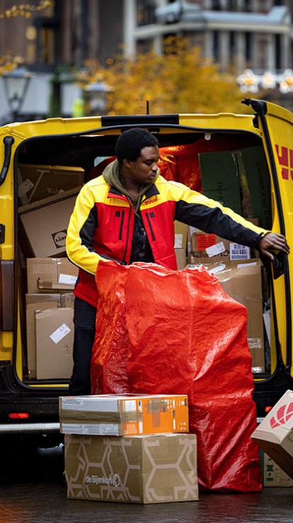 Pakketbezorger in werkkleding staat achter busje met stapel pakketten en een grote zak. Het busje staat midden op straat en is zelf ook gevuld met pakketten.