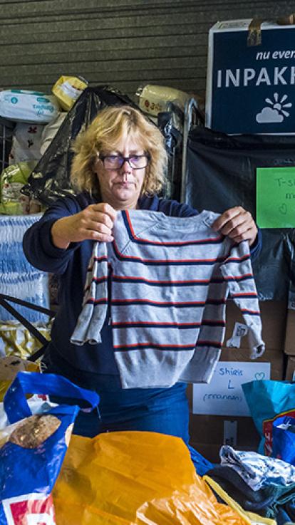 Vrouw houdt gestreept kindertruitje omhoog en bekijkt hem met een kritische blik. Ze is omringt met plastic zakken en karren met zakken en dozen erin.