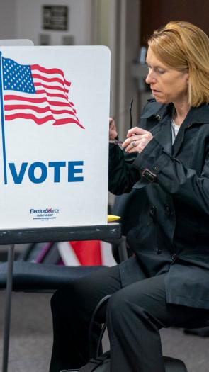Een vrouw zit op een stoel terwijl ze stemt voor de Amerikaanse verkiezingen. Voor haar een klein stemhokje op een tafel met de Amerikaanse vlag en 'Vote' erop.