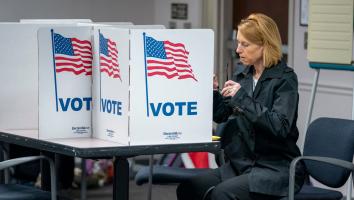 Een vrouw zit op een stoel terwijl ze stemt voor de Amerikaanse verkiezingen. Voor haar een klein stemhokje op een tafel met de Amerikaanse vlag en 'Vote' erop.