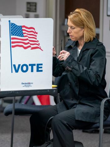 Een vrouw zit op een stoel terwijl ze stemt voor de Amerikaanse verkiezingen. Voor haar een klein stemhokje op een tafel met de Amerikaanse vlag en 'Vote' erop.