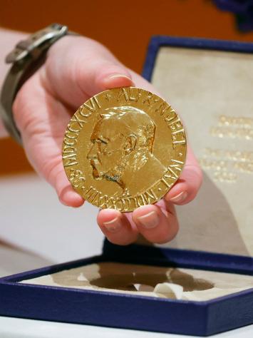 Een hand houdt een Nobelprijs voor de Vrede omhoog om te tonen aan de camera. De prijs zelf is een gouden cirkel met een portret van Nobel erop en tekst eromheen.