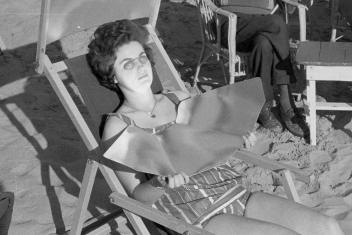 Zwart-witfoto van een vrouw die op een strandstoel ligt in haar badpak met een speciale bril en platen die de zon naar haar gezicht en hals weerspiegelen