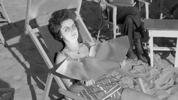 Zwart-witfoto van een vrouw die op een strandstoel ligt in haar badpak met een speciale bril en platen die de zon naar haar gezicht en hals weerspiegelen