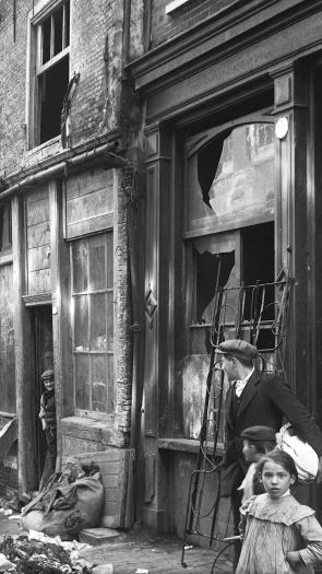Een zwart-witfoto van een arbeiderswijk begin 20ste eeuw in Amsterdam, met vervallen woningen, troep op straat en een aantal mensen die op straat lopen